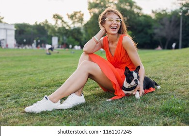 Happy Pretty Woman Sitting On Grass In Summer Park, Holding Boston Terrier Dog, Smiling Positive Mood, Wearing Orange Dress, Trendy Style, Slim Legs, Sneakers, Playing With Pet