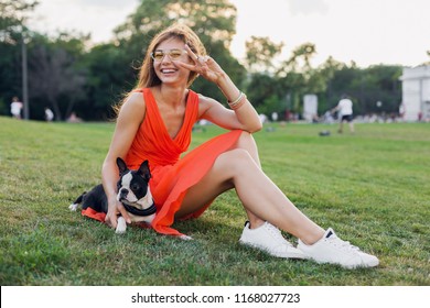 Happy Pretty Woman Sitting On Grass In Summer Park, Holding Boston Terrier Dog, Smiling Positive Mood, Wearing Orange Dress, Trendy Style, Slim Legs, Sneakers, Playing With Pet