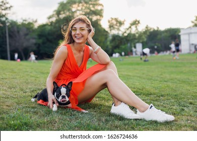 Happy Pretty Woman Sitting On Grass In Summer Park, Holding Boston Terrier Dog, Smiling Positive Mood, Wearing Orange Dress, Trendy Style, Slim Legs, Sneakers, Playing With Pet