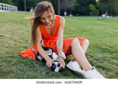 Happy Pretty Woman Sitting On Grass In Summer Park, Holding Boston Terrier Dog, Smiling Positive Mood, Wearing Orange Dress, Trendy Style, Slim Legs, Sneakers, Playing With Pet