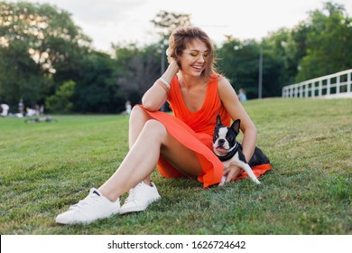 Happy Pretty Woman Sitting On Grass In Summer Park, Holding Boston Terrier Dog, Smiling Positive Mood, Wearing Orange Dress, Trendy Style, Slim Legs, Sneakers, Playing With Pet