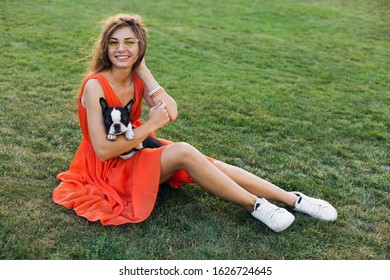 Happy Pretty Woman Sitting On Grass In Summer Park, Holding Boston Terrier Dog, Smiling Positive Mood, Wearing Orange Dress, Trendy Style, Slim Legs, Sneakers, Playing With Pet