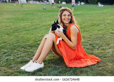 Happy Pretty Woman Sitting On Grass In Summer Park, Holding Boston Terrier Dog, Smiling Positive Mood, Wearing Orange Dress, Trendy Style, Slim Legs, Sneakers, Playing With Pet