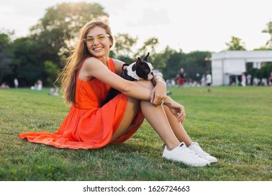 Happy Pretty Woman Sitting On Grass In Summer Park, Holding Boston Terrier Dog, Smiling Positive Mood, Wearing Orange Dress, Trendy Style, Slim Legs, Sneakers, Playing With Pet