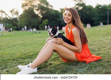 Happy Pretty Woman Sitting On Grass In Summer Park, Holding Boston Terrier Dog, Smiling Positive Mood, Wearing Orange Dress, Trendy Style, Slim Legs, Sneakers, Playing With Pet