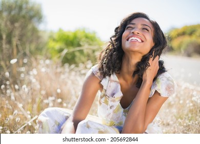 Happy Pretty Woman Sitting On The Grass In Floral Dress On A Sunny Day In The Countryside