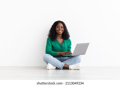 Happy pretty young black woman in casual outfit sitting on floor with modern laptop on white studio background, african american lady working online or surfing on Internet, copy space