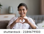 Happy pretty young Indian woman showing joined hands at chest, finger heart shape at camera, relaxing on comfortable sofa, enjoying home leisure, comfort. Blogger expressing love. Head shot portrait