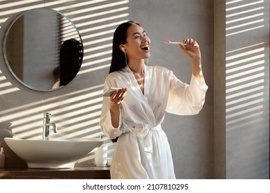 Happy Pretty Young Korean Woman In Bathrobe Singing While Brushing Teeth At Bathroom, Using Mobile Phone And Earphones, Listening To Music, Copy Space. Good Morning Concept