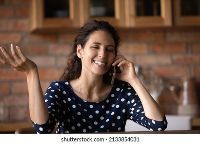 Happy Pretty Young Latin Woman Holding Distant Smartphone Call Conversation, Sharing Good News With Family Or Discussing Ideas With Friends, Sitting At Table In Kitchen, Distant Communication Concept.