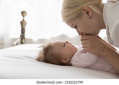 Happy Pretty Young Mom Cuddling Sweet Baby Lying On Back On Bed, Holding, Kissing Infant Arms. Devoted Mum Playing With Little Child With Love, Affection, Joy, Enjoying Motherhood, Family Time