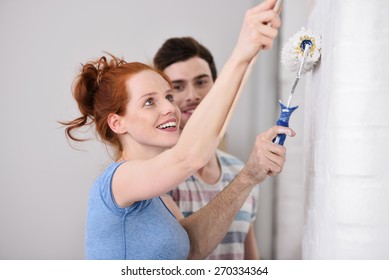 Happy Pretty Young Redhead Woman Painting A Wall With Her Husband Smiling As She Applies The Roller