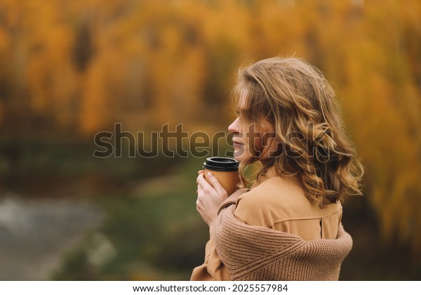 A happy pretty young woman in\
casual clothes enjoys solitude drinking coffee walking traveling by\
car in the autumn forest in nature in fall, selective\
focus