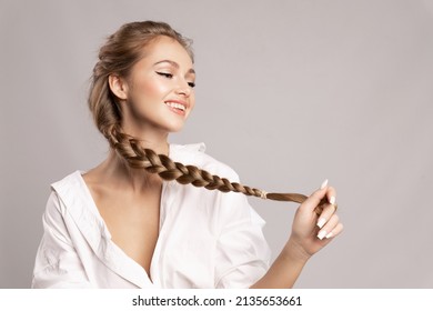 Happy Pretty Young Woman Holding Her Long Pigtail, Pleased With Healthy Shining Hairs Posing On Gray Background. Beauty Portrait Of Smiling Girl With Stylish Braid. Haircare Treatment Products Ad.