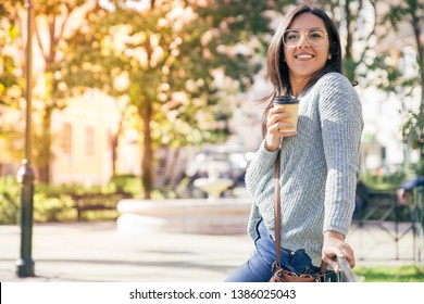 Happy pretty young woman sitting on railing outdoors. Beautiful lady holding drink in disposable cup with city view in background. Urban lifestyle concept. - Powered by Shutterstock