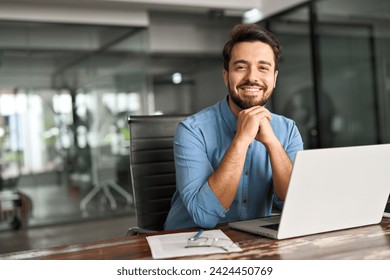 Happy professional business man company employee, young entrepreneur, smiling latin businessman working on laptop computer technology looking at camera working in office sitting at desk, portrait.