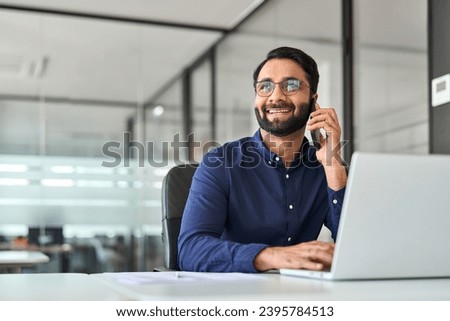 Happy professional busy Indian businessman talking on phone using laptop computer in office. Smiling business man making call on mobile cellphone, consulting client or having conversation at work.