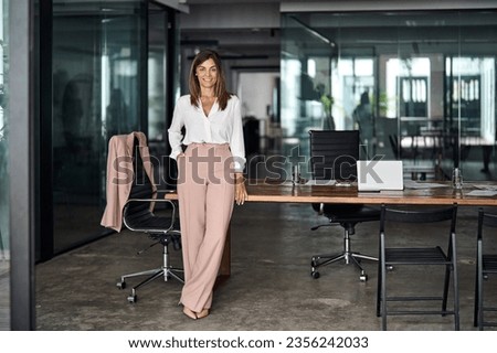Happy professional elegant mature 45 years old business woman, smiling Latin businesswoman executive manager leader, middle aged female entrepreneur standing at work in office. Full body portrait.