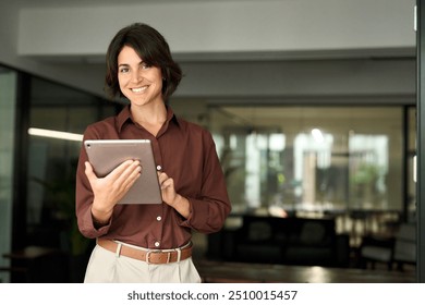 Happy professional female company executive, smiling Hispanic business woman entrepreneur, businesswoman corporate leader manager standing in office looking at camera using tablet. Portrait. - Powered by Shutterstock