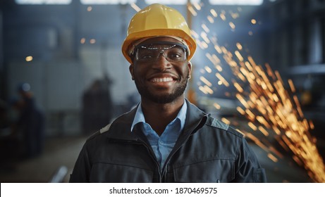 Happy Professional Heavy Industry Engineer Worker Wearing Uniform, Glasses And Hard Hat In A Steel Factory. Smiling African American Industrial Specialist Standing In A Metal Construction Manufacture.