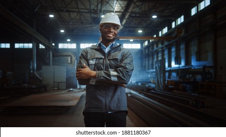 Happy Professional Heavy Industry Engineer Worker Wearing Uniform, Glasses And Hard Hat In A Steel Factory. Smiling African American Industrial Specialist Standing In A Metal Construction Manufacture.