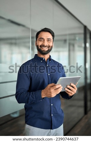 Happy professional Indian businessman using tab computer working standing in office. Smiling professional business man employee holding digital tablet tech device working, looking at camera. Vertical