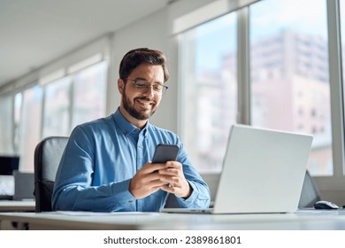 Happy professional latin business man company employee, smiling busy young businessman working on laptop holding smartphone using cellphone looking at mobile phone sitting at office workplace desk. - Powered by Shutterstock