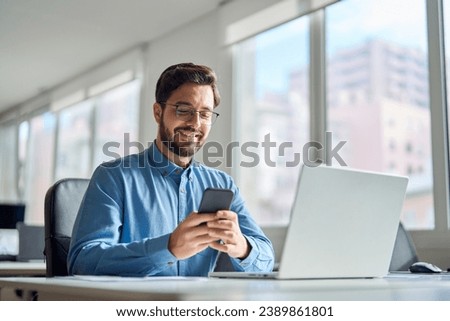 Happy professional latin business man company employee, smiling busy young businessman working on laptop holding smartphone using cellphone looking at mobile phone sitting at office workplace desk.