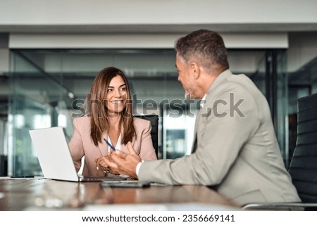 Happy professional mature Latin businessman and businesswoman executive colleagues wearing suits sitting at table having partnership business discussion using laptop at corporate meeting in office.