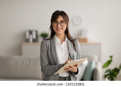 Happy professional middle eastern female psychologist writing in clipboard, looking and smiling at camera, working in modern office. Psychotherapy services, mental health professional concept - Shutterstock ID 2101132801