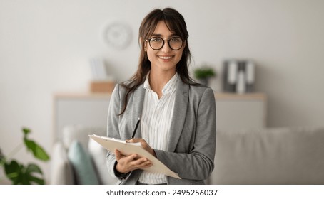 Happy professional middle eastern female psychologist writing in clipboard, looking and smiling at camera, working in modern office. Psychotherapy services, mental health professional concept - Powered by Shutterstock