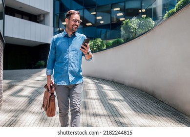 Happy professional wearing formals messaging on smart phone. Young businessman is carrying laptop bag. He is walking on footpath against office building in the city during sunny day. - Powered by Shutterstock