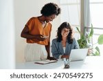 Happy professional women collaborating with enthusiasm in a modern office setting. Two female entrepreneurs using a laptop for a productive business meeting in a corporate workplace.