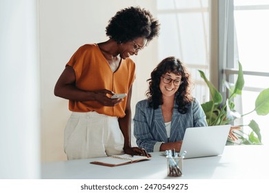 Happy professional women collaborating with enthusiasm in a modern office setting. Two female entrepreneurs using a laptop for a productive business meeting in a corporate workplace.