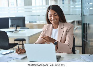 Happy professional young African American business woman manager executive wearing suit working in office elearning looking at laptop computer watching online webinar writing notes sitting at desk. - Powered by Shutterstock