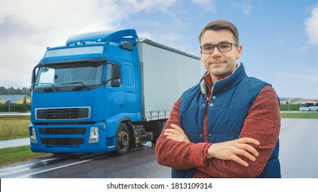 Happy Professional Young Truck Driver Crosses Arms And Smiles On Camera. Behind Him Parked Blue Long Haul Semi-Truck With Cargo Trailer