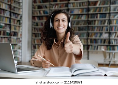 Happy Proud Gen Z Fresh College Girl In Headphones Showing Hand Like Sign, Thumb Up, Looking At Camera, Smiling, Working On Project In Library, Writing Essay, Research Paper Draft. Head Shot Portrait