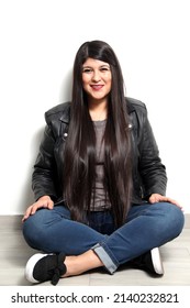 Happy And Proud Latin Young Adult Woman Shows Her Long Straight, Black And Shiny Hair On A White Background With A Leather Jacket And Black Tennis Shoes
