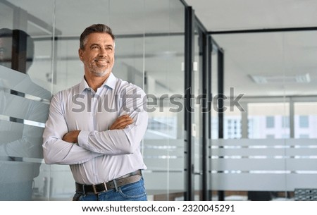 Happy proud mid aged older professional business man executive manager investor standing in office arms crossed leaning at glass wall looking away thinking of success, leadership, corporate growth.