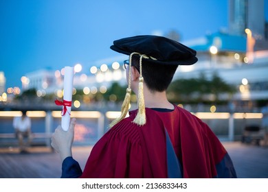 Happy Proud PhD Graduated Male Students In Academic Dress Gown With Certificate Facing Colorful City Nights. Back View