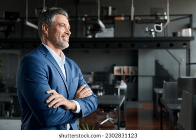 Happy proud prosperous mid aged mature professional business man ceo executive wearing suit standing in office arms crossed looking away thinking of success, leadership, side profile view.