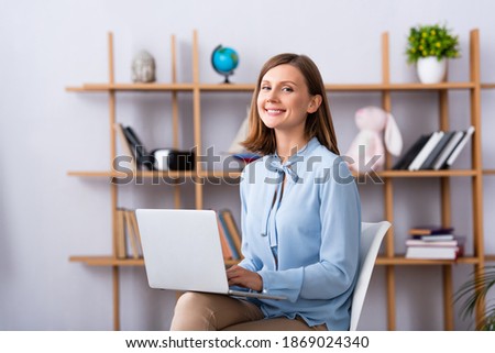 Happy psychologist looking at camera while using laptop in office on blurred background