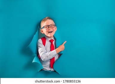 Happy Pupil In Uniform With Bag Breaking Through Blue Paper Wall. Funny Kid Boy With Red Backpack. Child Pointing Up On Text For Courses Secondary School Private.