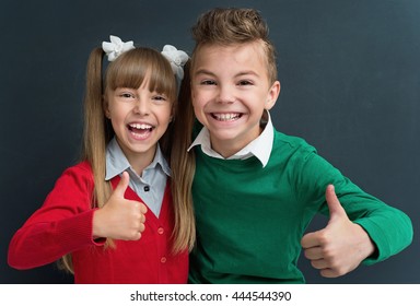 Happy Pupils - Boy And Girl, Showing Thumbs Up Gesture In Front Of A Big Chalkboard. Back To School Concept. 