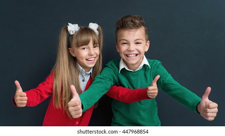 Happy Pupils - Boy And Girl, Showing Thumbs Up Gesture In Front Of A Big Chalkboard. Back To School Concept.