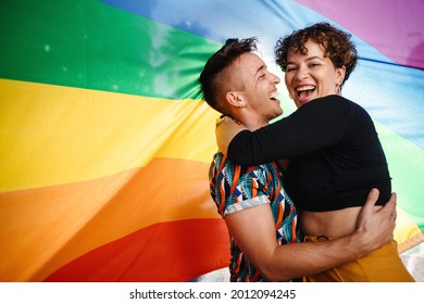 Happy queer couple standing against a rainbow pride flag. Young LGBTQ couple smiling cheerfully while embracing each other. Two non-conforming lovers celebrating gay pride together. - Powered by Shutterstock