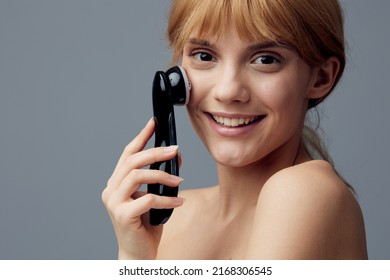 A Happy, Radiant Woman With Perfect Skin Stands On A Gray Background Wrapped In A Towel And Does A Facial Massage With A Black Electric Anti-aging Massager, Smiling Pleasantly At The Camera.