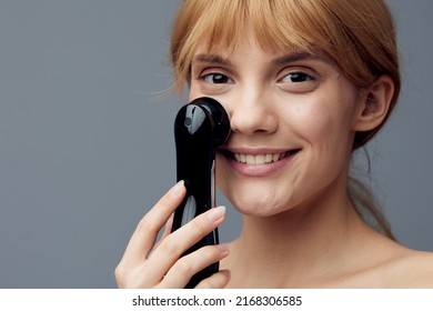 A Happy, Radiant Woman With Perfect Skin Stands On A Gray Background Wrapped In A Towel And Does A Facial Massage With A Black Electric Anti-aging Massager, Smiling Pleasantly At The Camera.