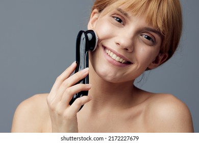 A Happy, Radiant Woman With Perfect Skin Stands On A Gray Background Wrapped In A Towel And Does A Facial Massage With A Black Electric Anti-aging Massager, Smiling Pleasantly At The Camera.