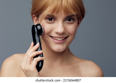 A Happy, Radiant Woman With Perfect Skin Stands On A Gray Background Wrapped In A Towel And Does A Facial Massage With A Black Electric Anti-aging Massager, Smiling Pleasantly At The Camera.